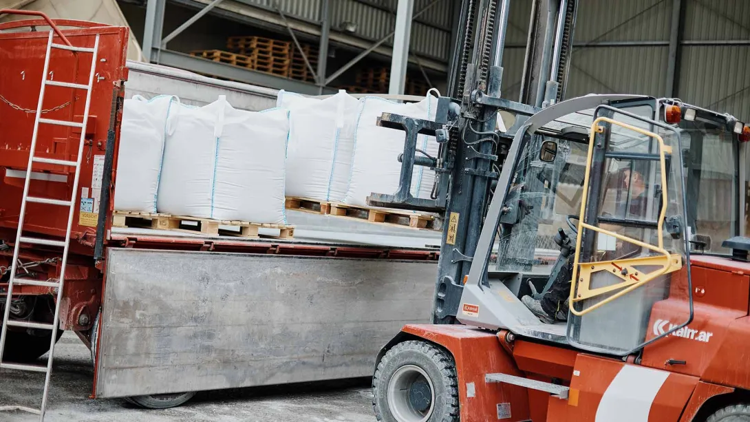 Ein Gabelstapler hebt große weiße Säcke, die auf einer Palette gestapelt sind, um sie in einen roten Lkw zu verladen. Der Hintergrund zeigt eine Lagerhalle, in der die Waren aufbewahrt werden.